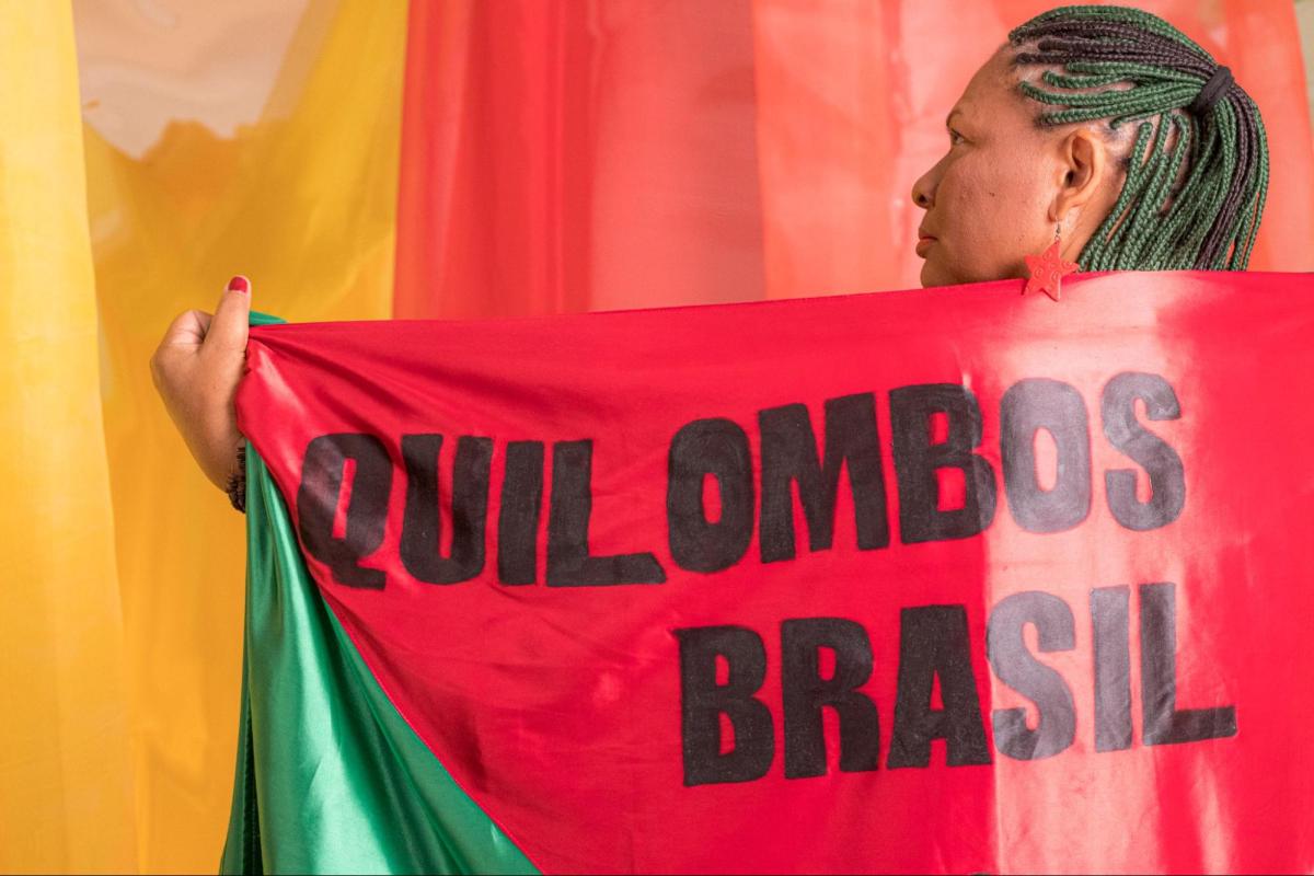 Ana Maria Eugênia da Silva, coordenadora estadual do Movimento Quilombola, do Quilombo Sitio Veiga, de Quixadá-CE, de costas para foto coberta com uma bandeira que diz: "Quilombos Brasil"